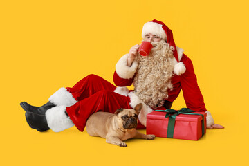 Santa Claus with Christmas gift, mug of tea and cute French bulldog on yellow background
