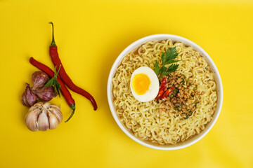 A bowl of instant noodles with egg, chili, garlic, and shallots on a vibrant yellow background. Perfect for Asian cuisine or cooking concept.