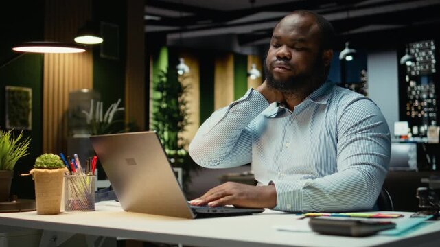 African american male worker stretching his back and neck muscles, correcting posture for muscle relief and relaxation. Strain and neck stiffness due to intense activity in office. Camera A.