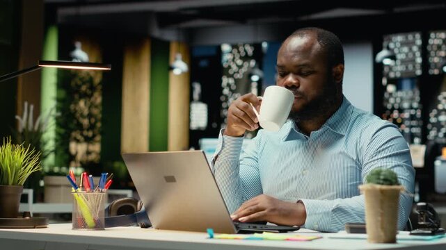 Black office employee serving a cup of coffee and doing overtime at workplace, staying awake for budgeting tasks in the strategy room after hours. Caffeine refreshment for proofreading. Camera B.