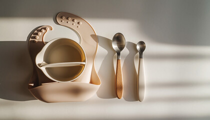 Modern baby feeding essentials a neutral-toned silicone bib, suction bowl, and child-friendly spoons arranged in natural light, perfect for infant and toddler meals