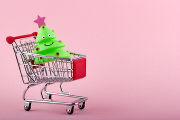 Grocery cart with small green toy Christmas tree on pink background. New Year shopping concept.