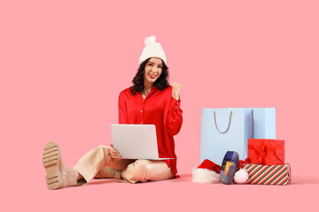 Pretty young woman with laptop pointing at shopping bags and Christmas gifts on pink background