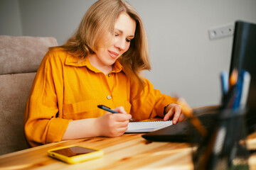 blonde woman in yellow shirt happily working in home office, writes notes and appears focused with...