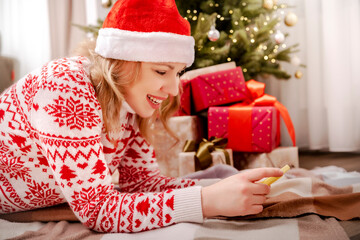 Blonde woman in Santa hat enjoys festive online chats with friends lying under Christmas tree surrounded by presents