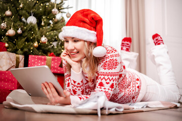 Blonde woman in Santa hat chatting online with friends on tablet under Christmas tree surrounded by gifts