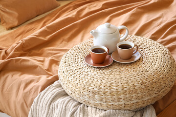 Wicker pouf with teapot and cups of tea on comfortable bed in interior of bedroom, closeup
