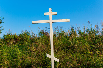 Prominent white Orthodox cross rising from vibrant green foliage against a bright blue sky, a symbol of devotion in nature's embrace. Russia