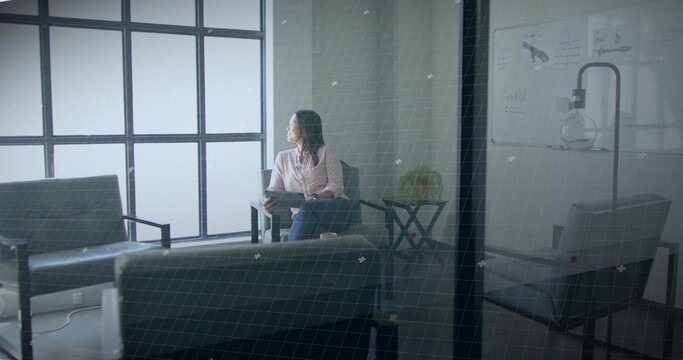 Observing Asian woman sitting thoughtfully in office lounge area, with tablet device and whiteboard