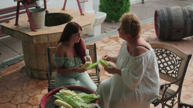 Mother and her daughter, enjoying the outdoors in a picturesque romanian village courtyard, husking fresh sweet corn side by side