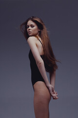 Fototapeta premium Young woman in black bodysuit posing against a neutral gray background with serious expression and long hair