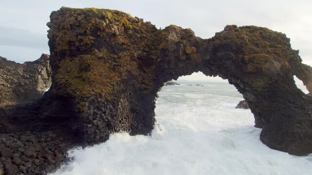 Aerial or drone footage of the stunning natural basalt rock arch of Gatklettur, also known as Hellnar Arch, located on the rugged coastline of Arnarstapi, Iceland.