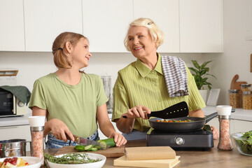 Happy senior woman frying vegetables while teaching her granddaughter cooking with recipe book in kitchen