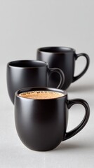 Three Black Ceramic Coffee Mugs With One Filled With Coffee And Cream Topping Arranged On A Textured Gray Surface With Soft Studio Lighting