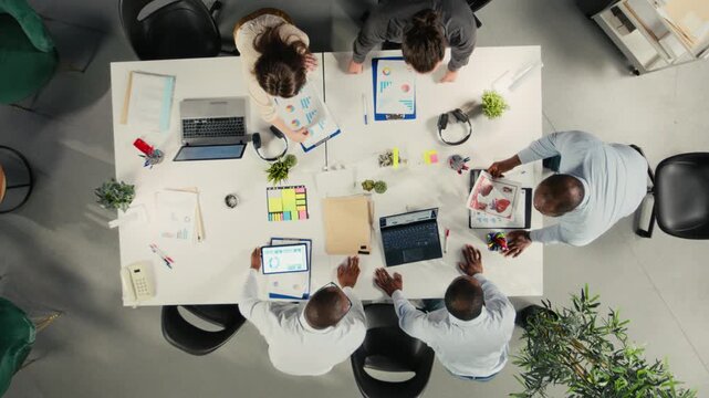 Top down view zoom in of employees discussing business intel and performance in meeting. Multinational company work team reviews reports, forecasts and diagrams to align strategy. Camera A.