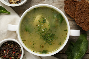 Bowl of tasty green borscht with spinach leaves, baguette slices and peppercorns on wooden background