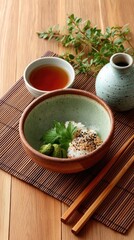 Top Down View Of A Ceramic Bowl Filled With White Rice Topped With Sesame Seeds And Fresh Green Herbs Accompanied By A Small Cup Of Tea And A Small Decorative Vase On A Wooden Table