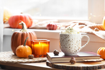 Cup of pumpkin latte with book and autumn decor on table near window at home