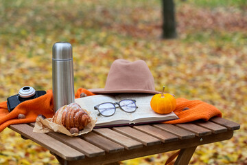 Book with eyeglasses, croissant and thermos on table in autumn park