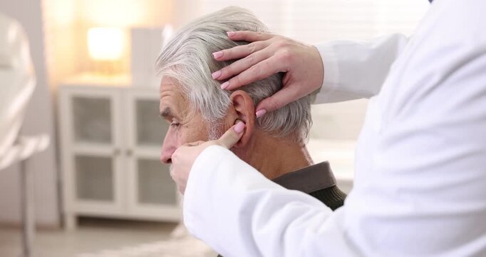 Doctor putting hearing aid into senior man's ear in hospital, closeup