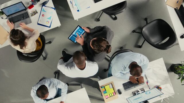 Top down view zoom out of work team in multinational company analyzing metrics and projections. Staff collaborates on strategy planning and report sharing to support business targets. Camera A.