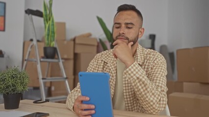 Man holding blue tablet with hand on chin in building surrounded by moving boxes and ladder; moving planning concern.