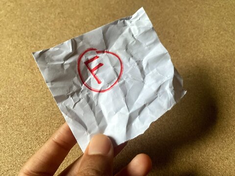 A person is holding a small, crumpled piece of paper with the failing grade "F" circled in red. Represents failure, disappointment, poor results, and academic struggle.