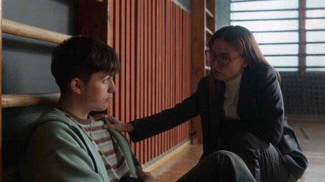 Young female school psychologist coming up to upset teenage boy, putting her hand on his shoulder, looking at him and telling him that he may share his trouble with her
