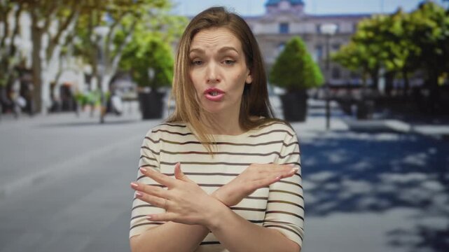 Young caucasian woman crossing forearms in sunlit city street with potted plants in background; rejection.