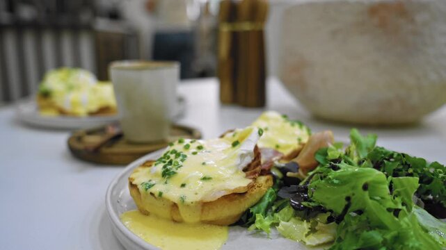 Brunch setting featuring eggs benedict and salad on a white table at a cafeteria with a blurred background enhancing the cozy dining ambiance