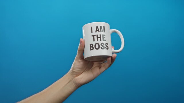 Hand holding white mug with bold text 'i am the boss' against solid blue wall background, symbolizing confidence and authority, ideal for corporate or leadership themes.