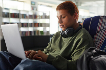 Young man sitting in modern library using laptop, wearing headphones around neck, focusing on typing, studying or working on digital project in relaxed environment