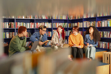 Group of multiethnic young adults sitting in library discussing ideas and taking notes, diverse men and women engaging in collaborative study session surrounded by bookshelves