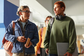 Multiethnic group of teenagers walking in hallway, Asian male holding books and talking to biracial male carrying laptop and wearing headphones, female teenager in background