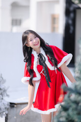 young asian woman wearing a red santa dress with white fur trim smiling and posing outdoors in a snowy Christmas scene, surrounded by frosted pine trees and bright winter sunlight