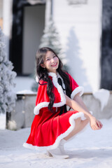 young asian woman wearing a red santa dress with white fur trim smiling and posing outdoors in a snowy Christmas scene, surrounded by frosted pine trees and bright winter sunlight
