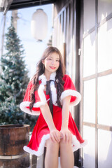 young asian woman wearing a red santa dress with white fur trim smiling and posing outdoors in a snowy Christmas scene, surrounded by frosted pine trees and bright winter sunlight