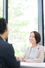 新緑の窓際で保険や企画の営業をするスーツの女性、会社で商談や打ち合わせするビジネスマンの男女2人