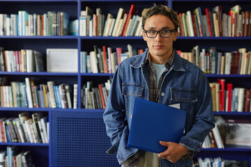 Portrait of Asian young adult man with short hair and glasses standing in front of bookshelf holding blue folder, looking directly at camera in library setting © Seventyfour