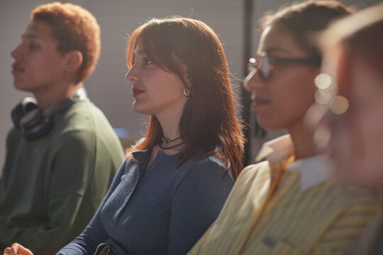 Diverse group of young adults and teenagers sitting in row, listening attentively during seminar or lecture, focus on Caucasian young woman with brown hair in center of frame - Powered by Adobe