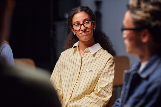 Young adult oman with curly hair and glasses smiling while listening to conversation in group setting, sitting among peers, engaging in discussion