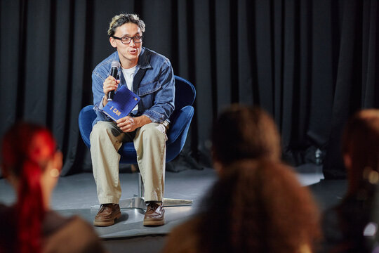 Young adult man sitting on stage holding microphone and notebook, speaking to audience during event, wearing glasses, engaging with group of people in front of him