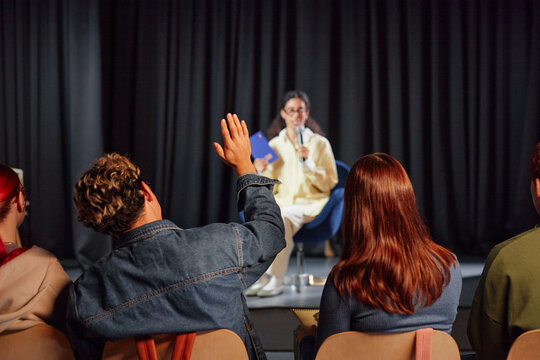 Young adult woman sitting on stage speaking into microphone while multiethnic group of young adults sitting in audience, listening with one raising hand to ask question