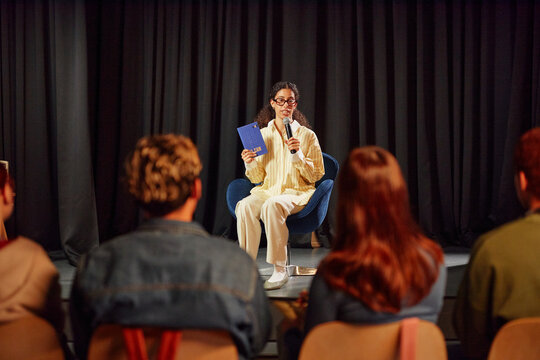 Young adult woman sitting on stage holding microphone and book, speaking to audience of young adults, engaging in public presentation or discussion event - Powered by Adobe