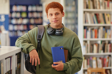 Portrait of teenage boy standing in library holding notebook and tablet, wearing headphones around neck, looking into camera with relaxed expression, carrying backpack