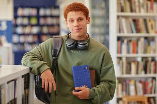 Portrait of teenage boy standing in library holding notebook and tablet, wearing headphones around neck, looking into camera with relaxed expression, carrying backpack - Powered by Adobe