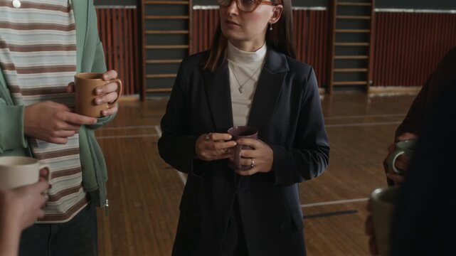 Tilt down of young female psychologist and teenagers with cups of tea or coffee discussing main points of forthcoming session while standing in circle in sports hall