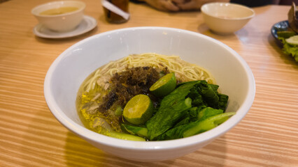 A bowl of Asian noodle soup with green vegetables, calamansi, shredded meat, and savory broth served on a wooden table, showcasing traditional comfort food cuisine