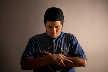 A Muslim man in traditional attire and cap performs the Qiyam (standing) posture of prayer (Shalat), with hands crossed over the chest. Dramatic lighting conveys deep spiritual focus
