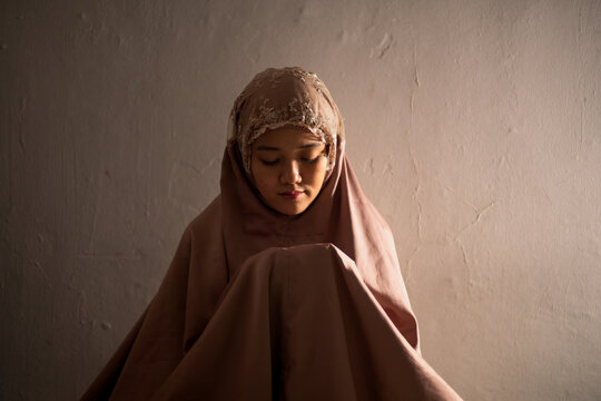 A Muslim woman in a dusty rose colored mukena performs prayer (shalat/salat) in a solemn, dimly lit room. The dramatic side lighting highlights the moment of spiritual devotion